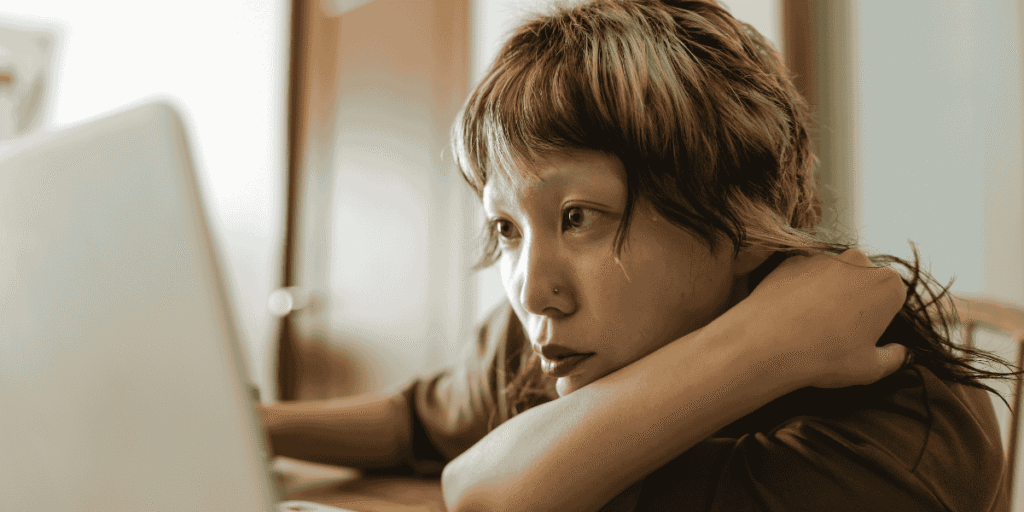 Junge Frau sitzt vor einem Laptop, wirkt erschöpft und stützt ihren Kopf auf den Arm, während sie auf den Bildschirm schaut