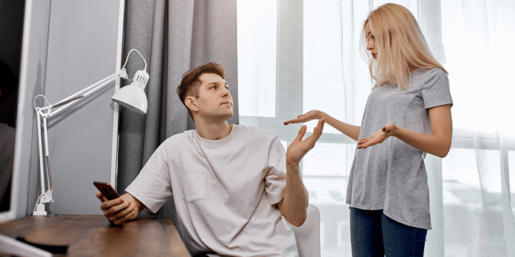 Zwei Personen im Raum gestikulieren einander gegenüber; eine sitzt am Schreibtisch mit Smartphone, die andere steht.