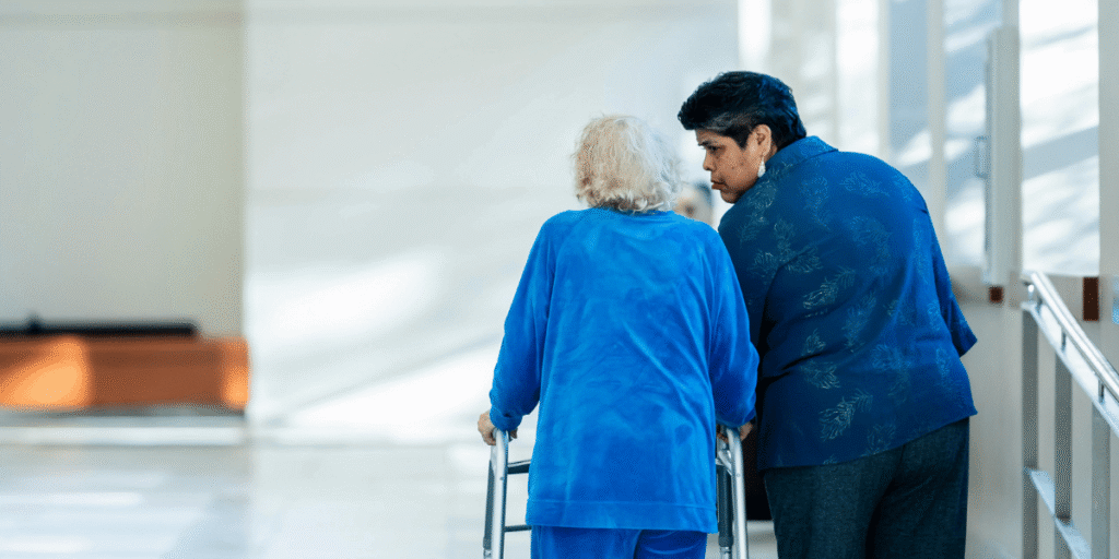 Zwei Personen von hinten im Flur: ältere Person mit Rollator wird von Begleitperson gestützt, beide in blauer Kleidung, Innenraum mit Geländer und Tageslicht.