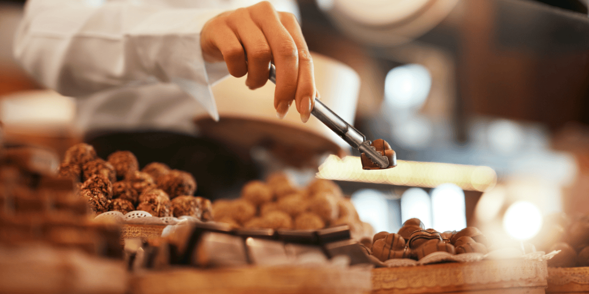 Hand mit Zange greift Praline aus Auslage mit verschiedenen Schokoladentrüffeln in Konditorei.