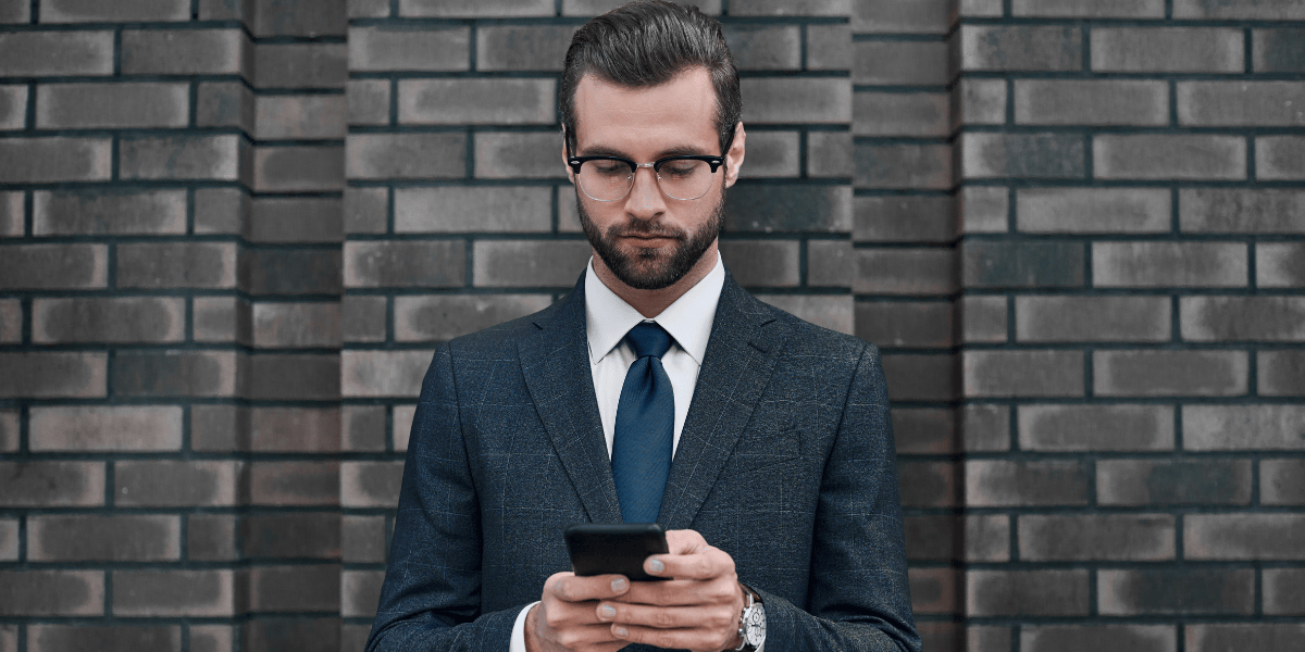 Person im Anzug mit Brille blickt auf ein Smartphone vor einer dunklen Ziegelwand