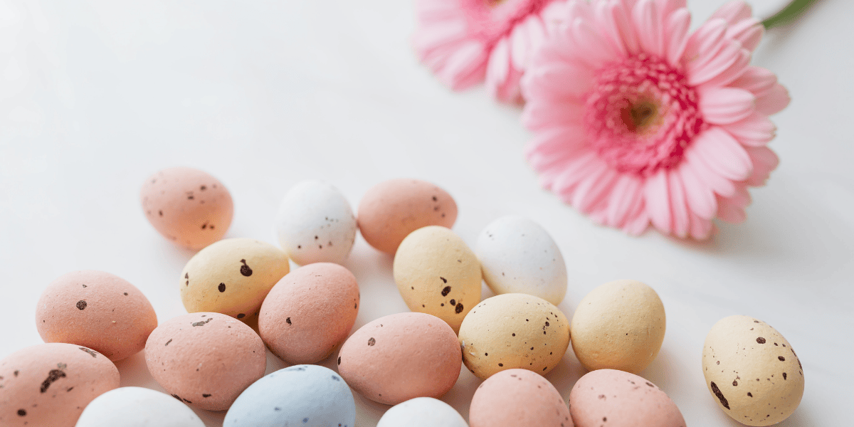 Pastellfarbene, gesprenkelte Schokoladeneier neben rosa Gerbera-Blüten auf hellem Hintergrund