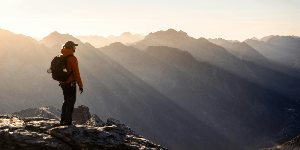Person mit Rucksack steht auf felsigem Gipfel und blickt über Berglandschaft im Sonnenlicht mit langen Schatten.