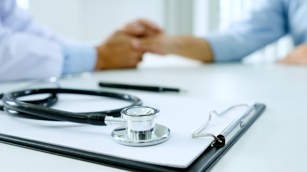 Stethoskop und Klemmbrett liegen auf einem Tisch, während im Hintergrund Arzt und Patient sich die Hände halten