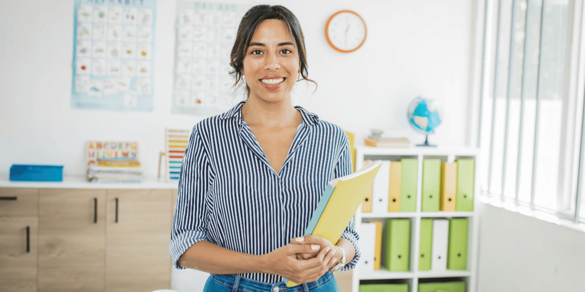Lehrerin im Klassenzimmer hält Bücher, steht vor Regal mit Ordnern, Weltkarte und Uhr im Hintergrund
