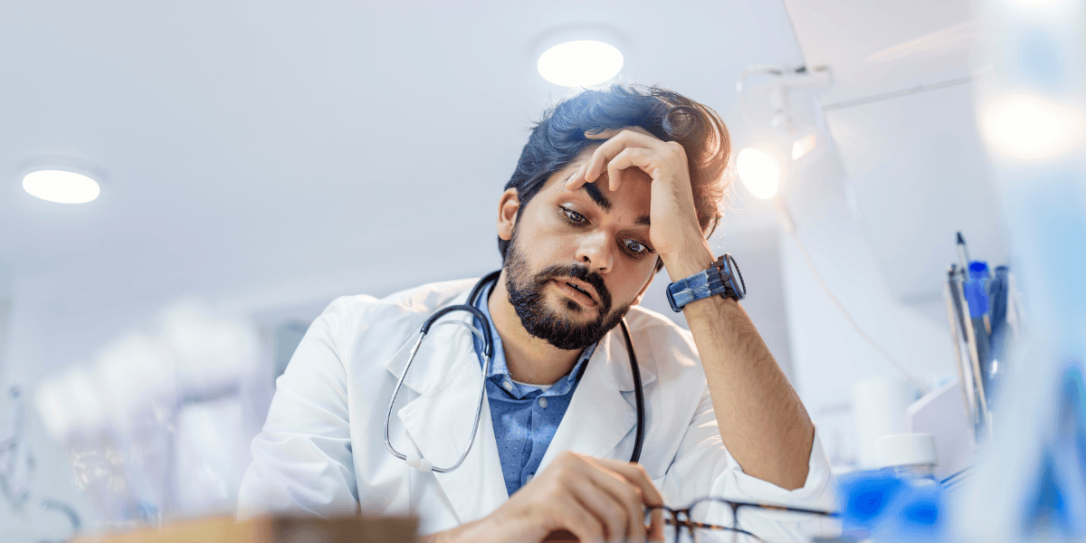 Arzt im Kittel mit Stethoskop sitzt am Schreibtisch, stützt den Kopf mit der Hand und blickt auf eine Brille; Klinikraum im Hintergrund.