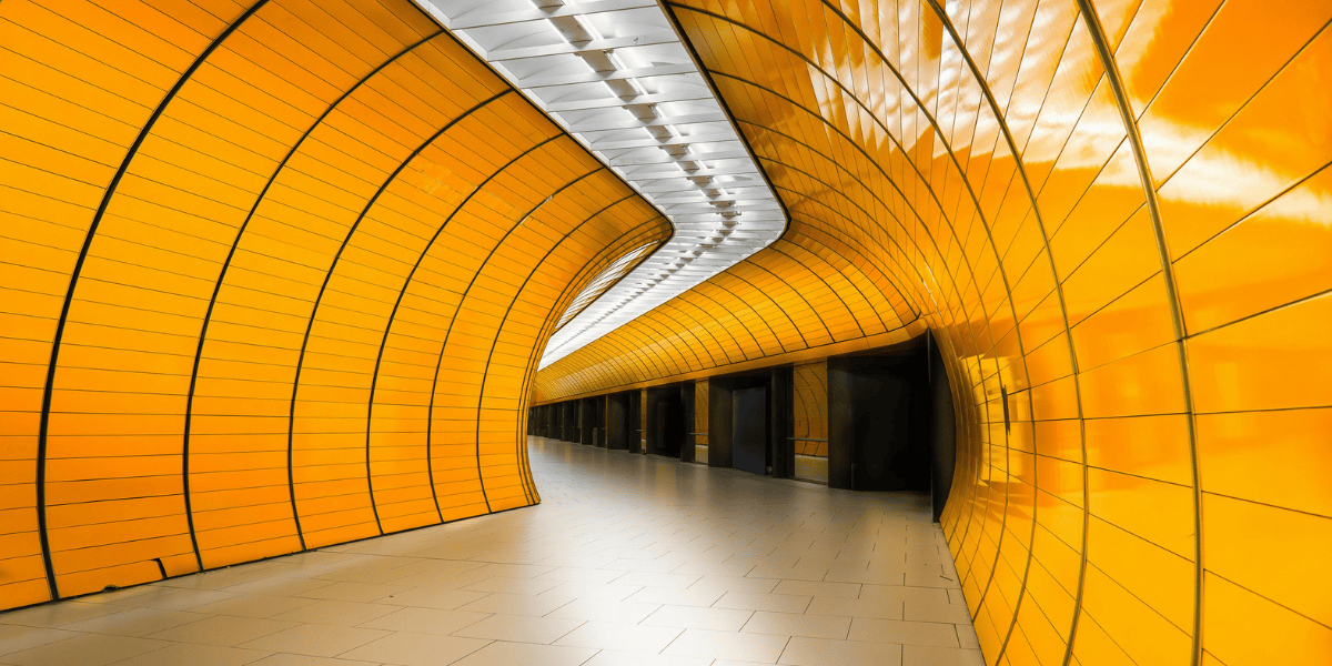 Geschwungener Tunnel mit orange beleuchteten Wandpaneelen, heller Boden und Deckenlichtstreifen, menschenleer.