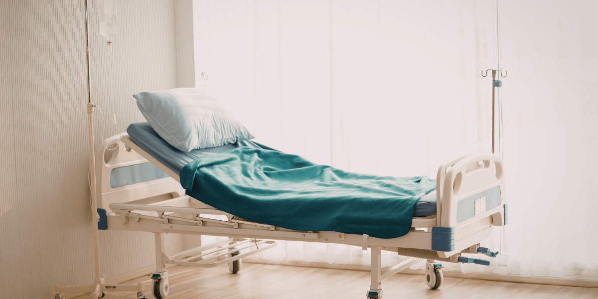 Leeres Krankenhausbett mit Kissen, Decke und Infusionsständer am Fenster.