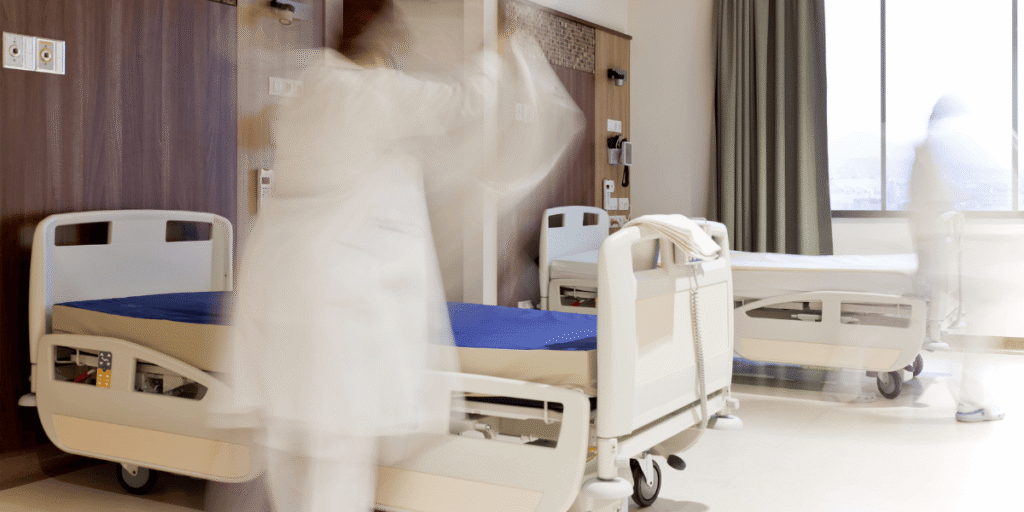 Krankenhauszimmer mit zwei Betten; unscharfe Person in Bewegung zwischen den Betten.