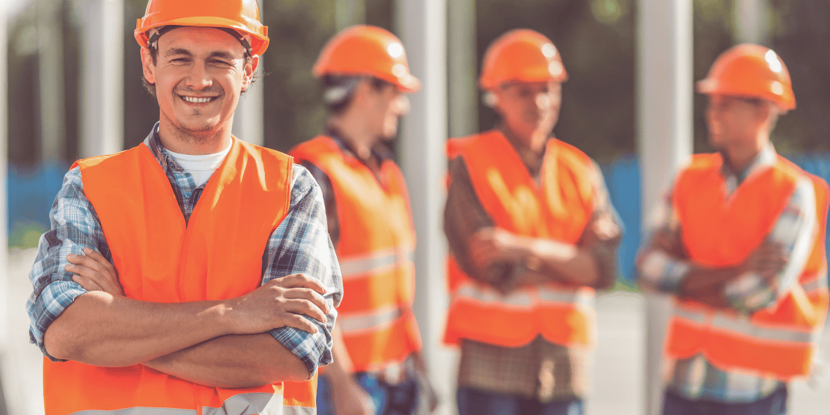 Arbeiter in orangefarbener Warnweste und Schutzhelm steht mit verschränkten Armen im Vordergrund; drei weitere Arbeiter in ähnlicher Schutzkleidung unscharf im Hintergrund auf einer Baustelle.