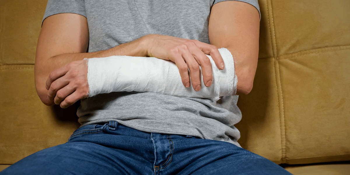 Person sitzt auf Sofa und hält einen Unterarm mit weißem Gipsverband; graues T-Shirt und Jeans sichtbar.