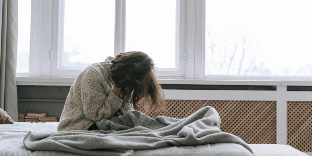 Person sitzt auf einem Bett vor einem Fenster, hat den Kopf in die Hände gelegt und wirkt erschöpft oder belastet