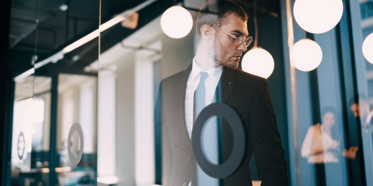 Person im Anzug mit Brille steht hinter Glaswand im Büro; runde Deckenleuchten und Reflexionen sichtbar.