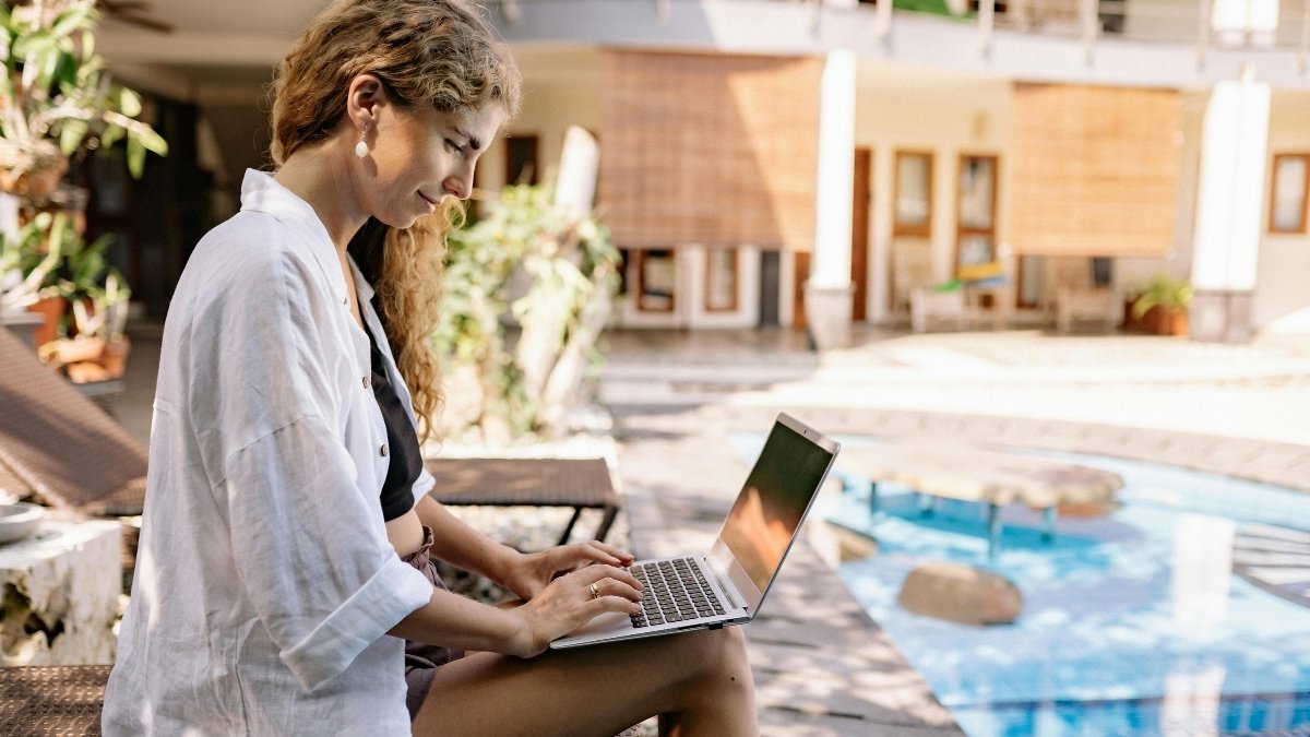 Junge Frau sitzt entspannt am Pool und arbeitet mit einem Laptop in sonniger Urlaubsatmosphäre.