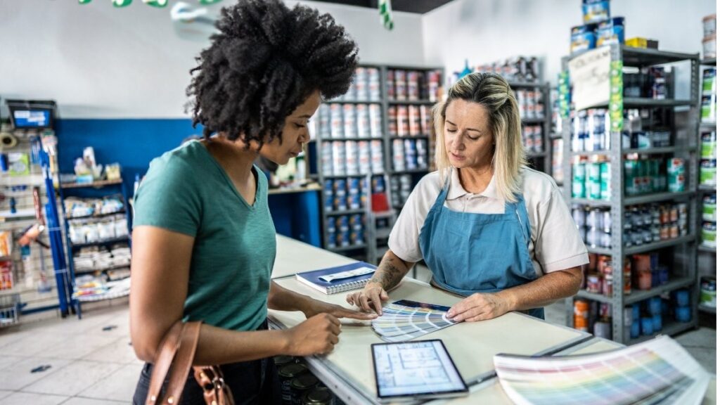 Zwei Frauen stehen in einem Geschäft an der Theke und besprechen gemeinsam Farbmuster, während im Hintergrund Regale mit Waren zu sehen sind