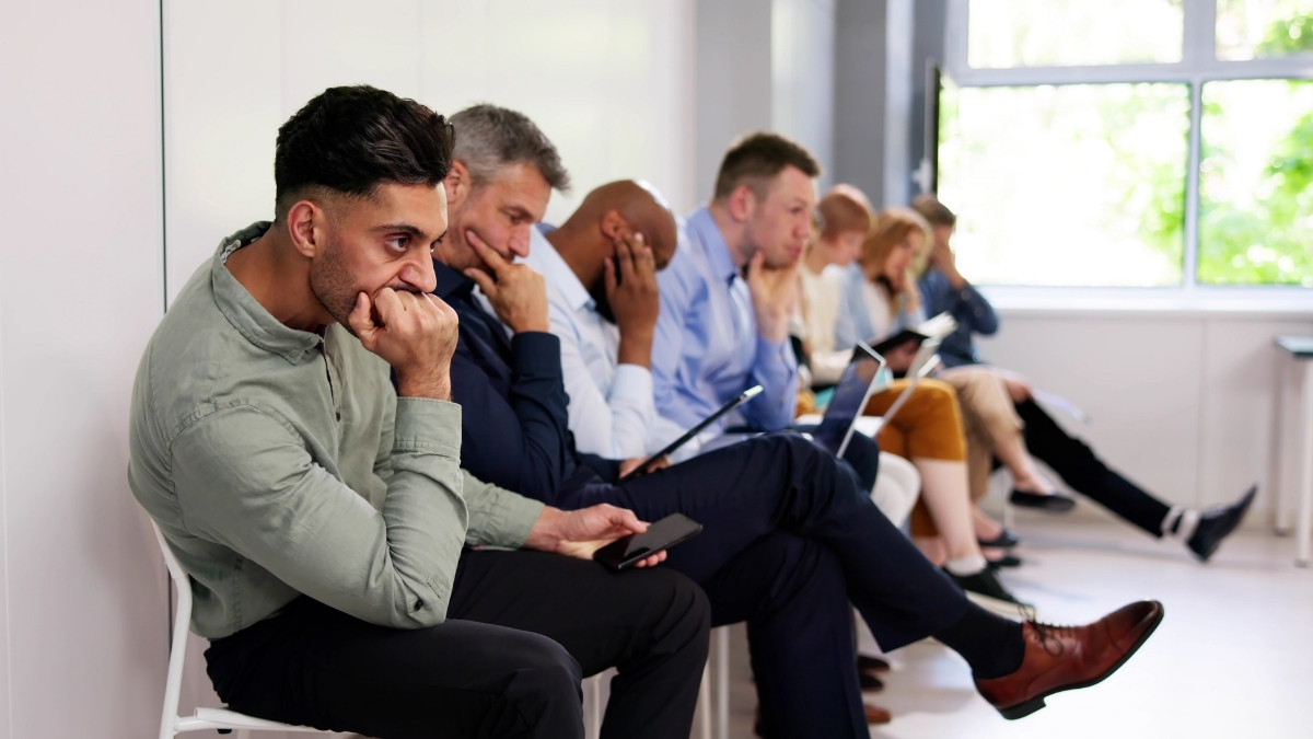 Mehrere Menschen sitzen nebeneinander in einem Wartebereich, schauen angespannt auf ihre Smartphones oder Unterlagen und wirken nachdenklich.