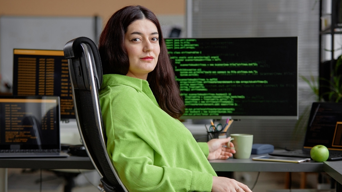 Junge Frau sitzt vor mehreren Bildschirmen mit Programmcode und blickt selbstbewusst in die Kamera.