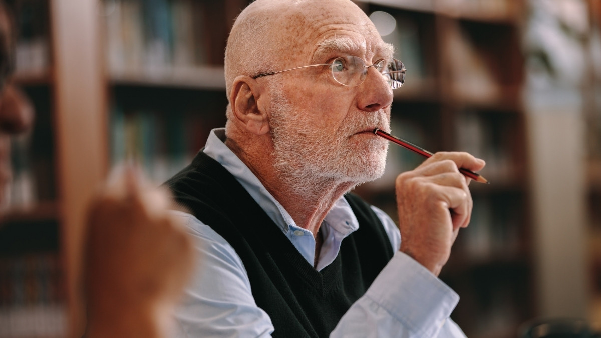 Älterer Mann mit Brille hält einen Stift an die Lippen und blickt nachdenklich zur Seite, sitzt in einer Umgebung mit Bücherregalen