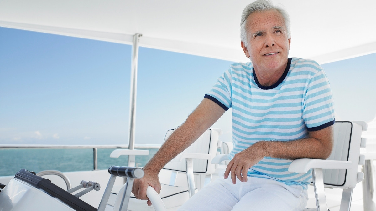 Älterer Mann sitzt entspannt auf dem Deck eines Bootes, hält das Steuerrad und blickt zufrieden in die Ferne über das Meer
