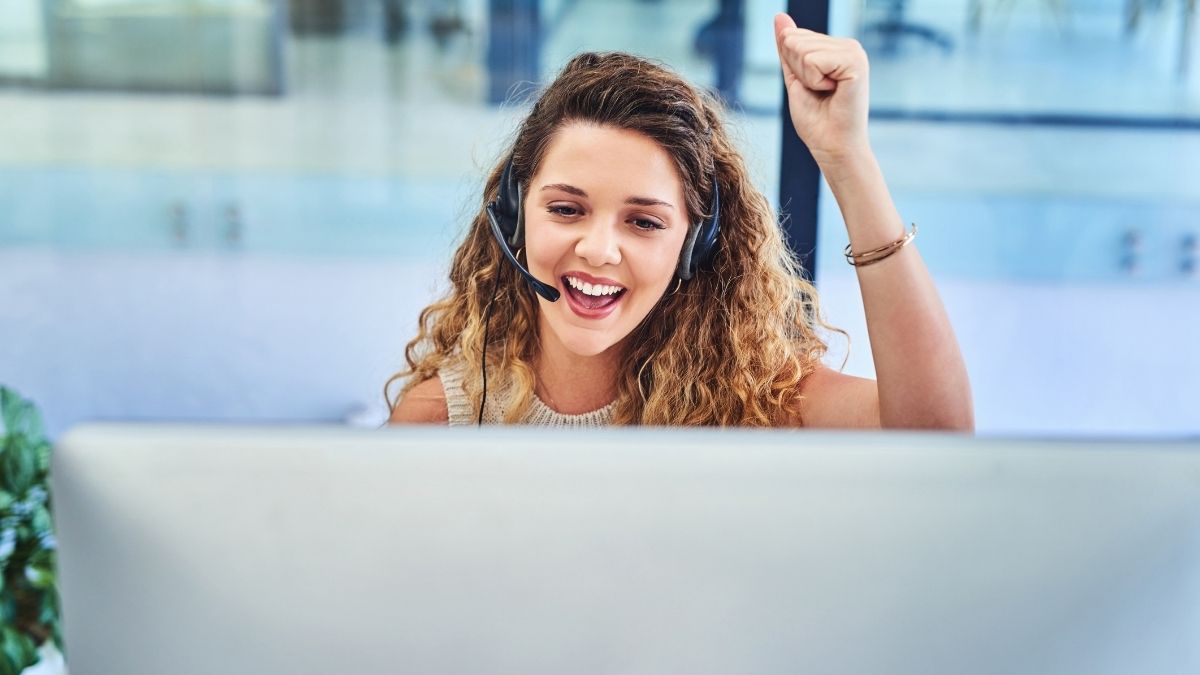 Lächelnde Frau mit Headset sitzt vor einem Computer und hebt die Faust