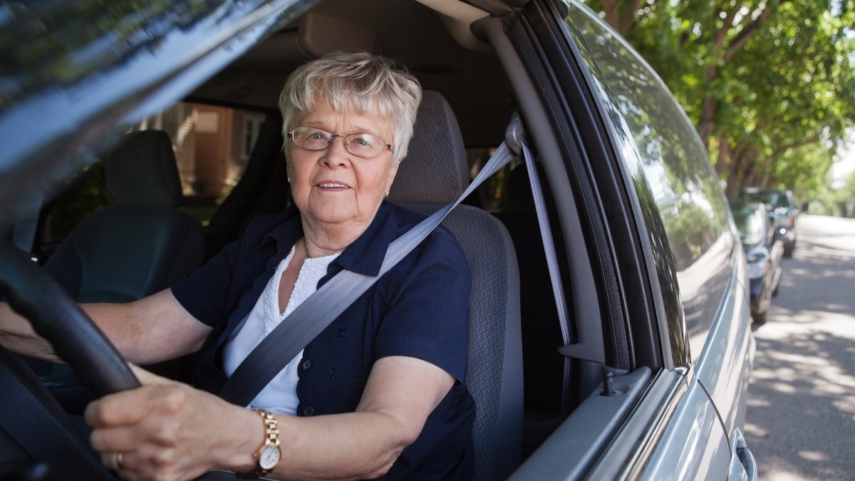 Ältere Frau mit Brille sitzt angeschnallt am Steuer eines Autos und blickt aus dem geöffneten Seitenfenster.