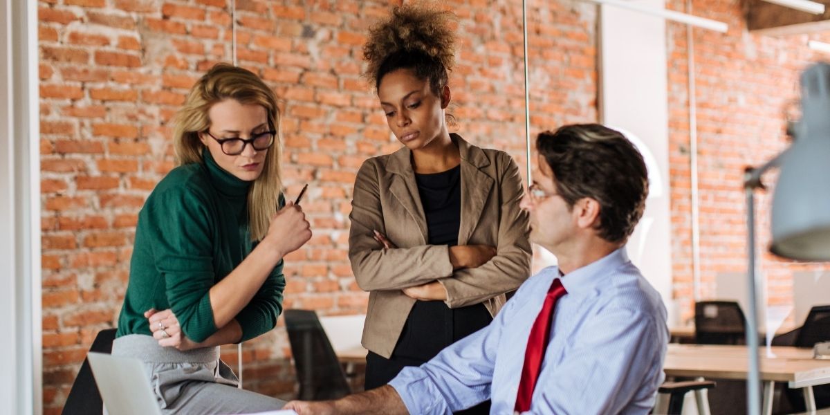 Drei Personen besprechen Dokumente in einem modernen Büro mit Backsteinwand und Computerarbeitsplatz.