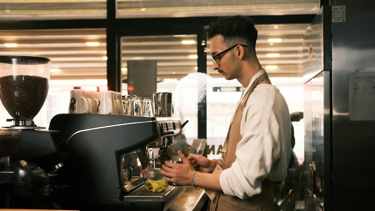 Ein Barista arbeitet an einer Kaffeemaschine hinter dem Tresen eines Cafés und bereitet ein Getränk zu.