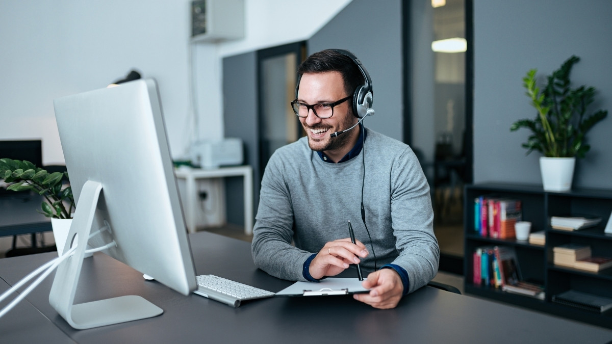Lächelnder Mann mit Headset sitzt vor einem Computer im Büro und macht sich Notizen während eines Gesprächs.