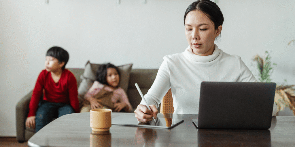 Frau arbeitet am Tisch mit Laptop und Tablet, schreibt mit Stift; zwei Kinder sitzen unscharf im Hintergrund auf einem Sofa.