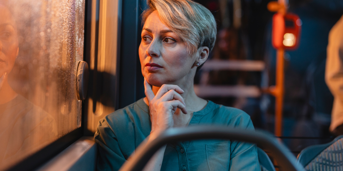 Frau mit kurzen Haaren sitzt am Fenster eines Busses, Hand am Kinn, Blick zur Seite, warme Innenbeleuchtung und verschwommene Stadtlichter im Hintergrund.