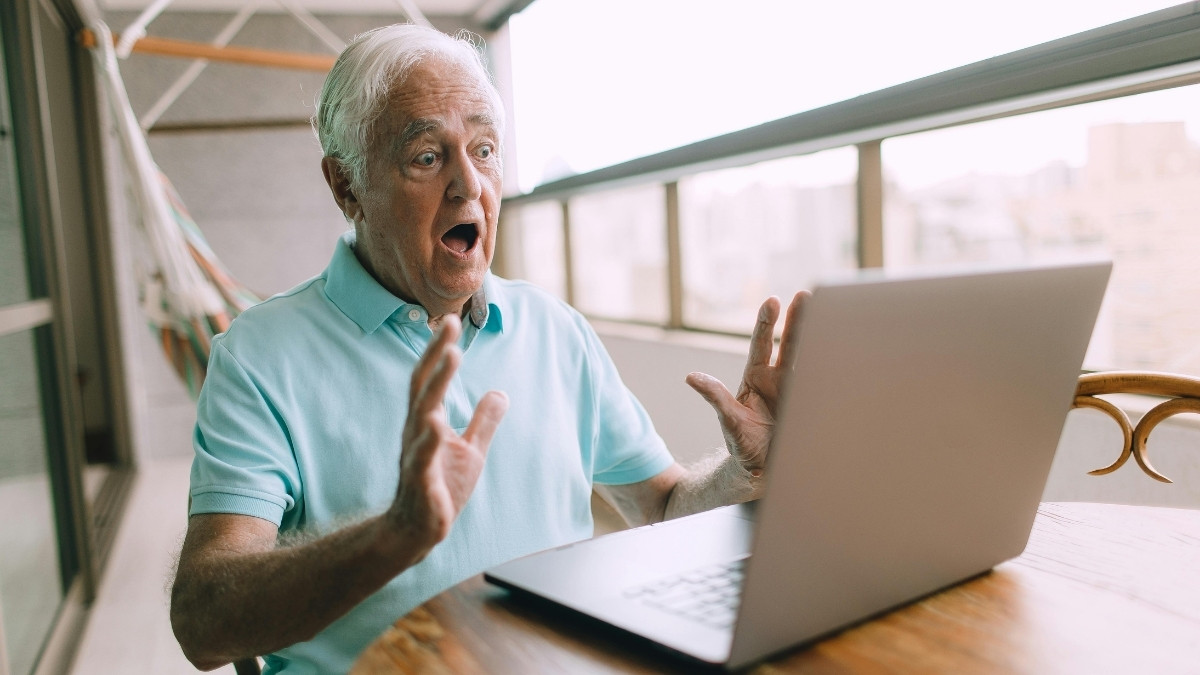 Überraschter älterer Mann sitzt auf einem Balkon vor einem Laptop und reagiert mit weit aufgerissenen Augen und erhobenen Händen auf den Bildschirm.