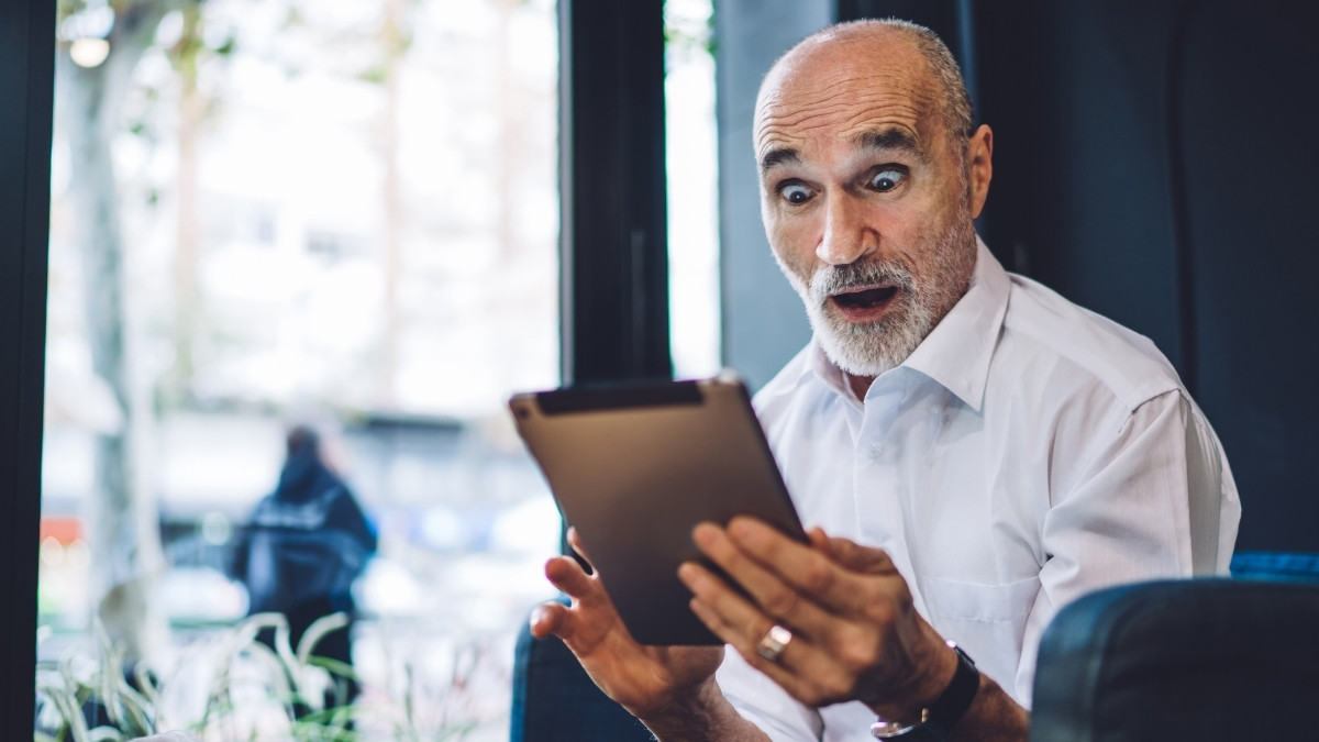 Ein älterer Mann schaut überrascht auf ein Tablet, das er in den Händen hält, und reagiert mit weit geöffneten Augen und offenem Mund.