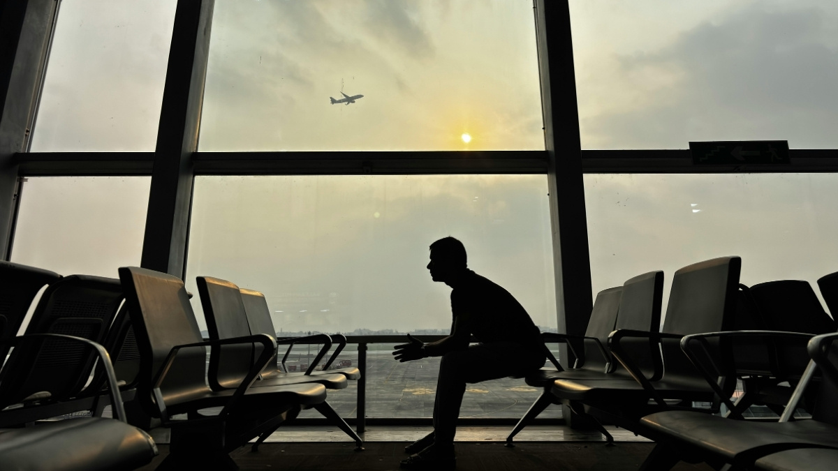 Silhouette einer sitzenden Person in einem Flughafen-Wartebereich vor großen Fenstern, während draußen ein Flugzeug am Himmel zu sehen ist.