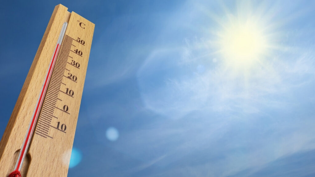 Thermometer zeigt hohe Temperatur vor blauem Himmel und strahlender Sonne