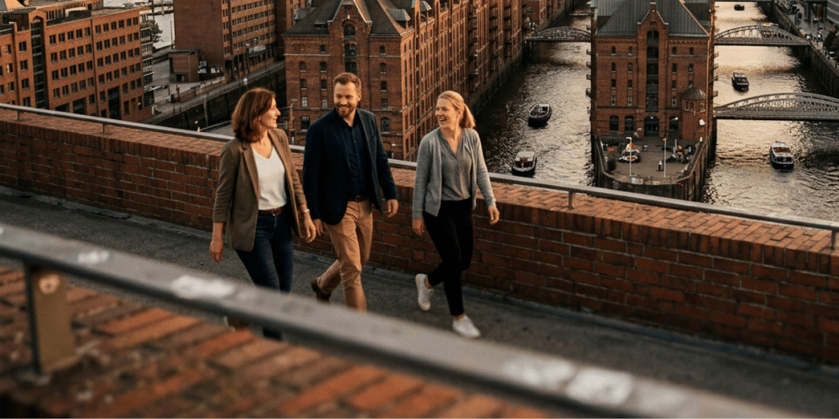 Drei Personen gehen auf einer erhöhten Promenade aus Backstein entlang eines Kanals mit historischen Lagerhäusern und kleinen Booten im Wasser.