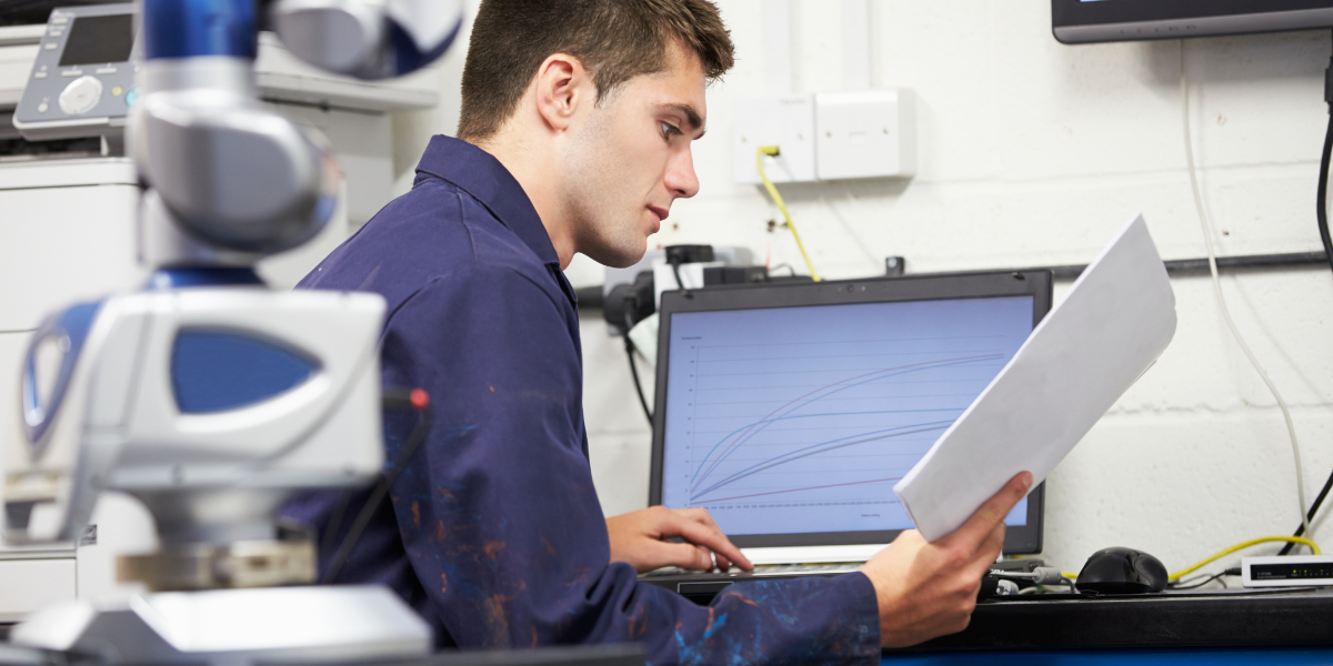 Person in Arbeitskleidung sitzt am Schreibtisch, hält ein Dokument und blickt auf Laptop mit Diagramm; Laborgerät im Vordergrund.