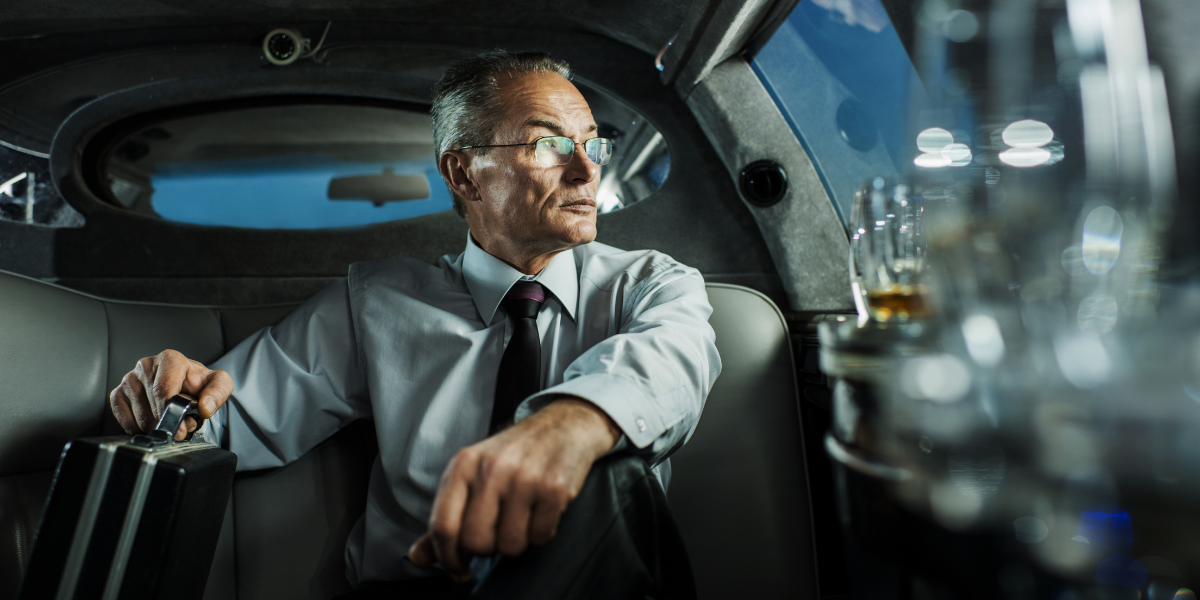 Älterer Mann mit Brille und Aktenkoffer sitzt in einer Limousine und blickt aus dem Fenster.