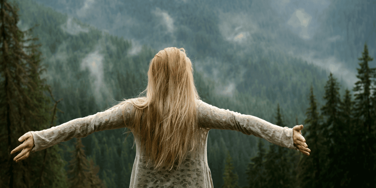 Person mit langen Haaren und ausgebreiteten Armen vor bewaldeter Berglandschaft im Nebel, von hinten fotografiert.