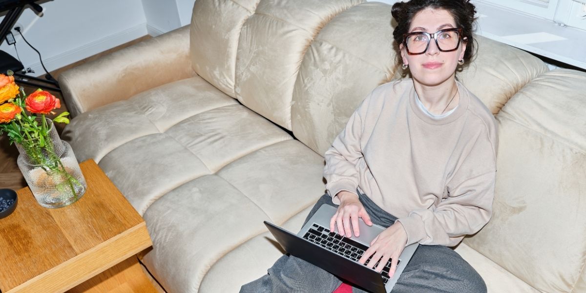 Person mit Brille arbeitet zu Hause an einem Laptop auf einem Sofa.