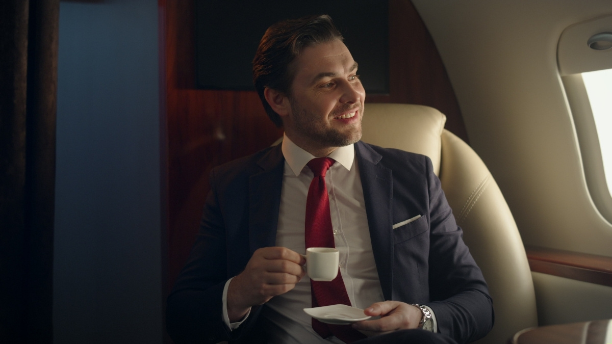 Ein Geschäftsmann im Anzug sitzt entspannt in einer luxuriösen Flugzeugkabine, trinkt Kaffee und blickt lächelnd aus dem Fenster.