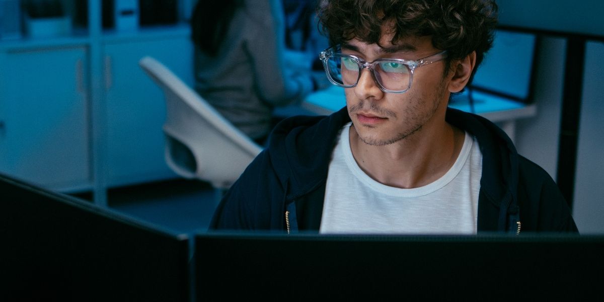 Person mit Brille sitzt vor mehreren Monitoren in einem Büro und blickt auf den Bildschirm; weitere Person unscharf im Hintergrund.