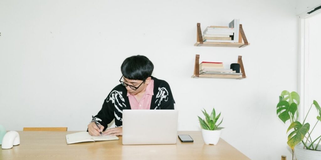 Person schreibt an einem Tisch neben Laptop und Smartphone in ein Notizbuch.