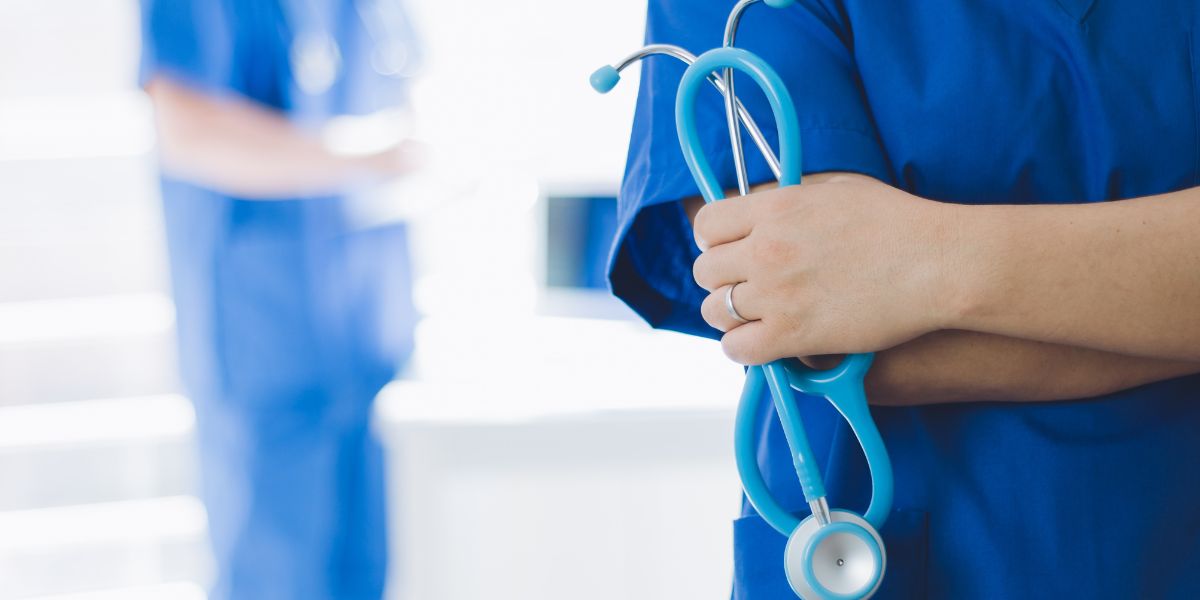 Pflegekraft in blauer Arbeitskleidung hält ein Stethoskop vor dem Oberkörper, unscharfe medizinische Fachkraft im Hintergrund einer Klinik.