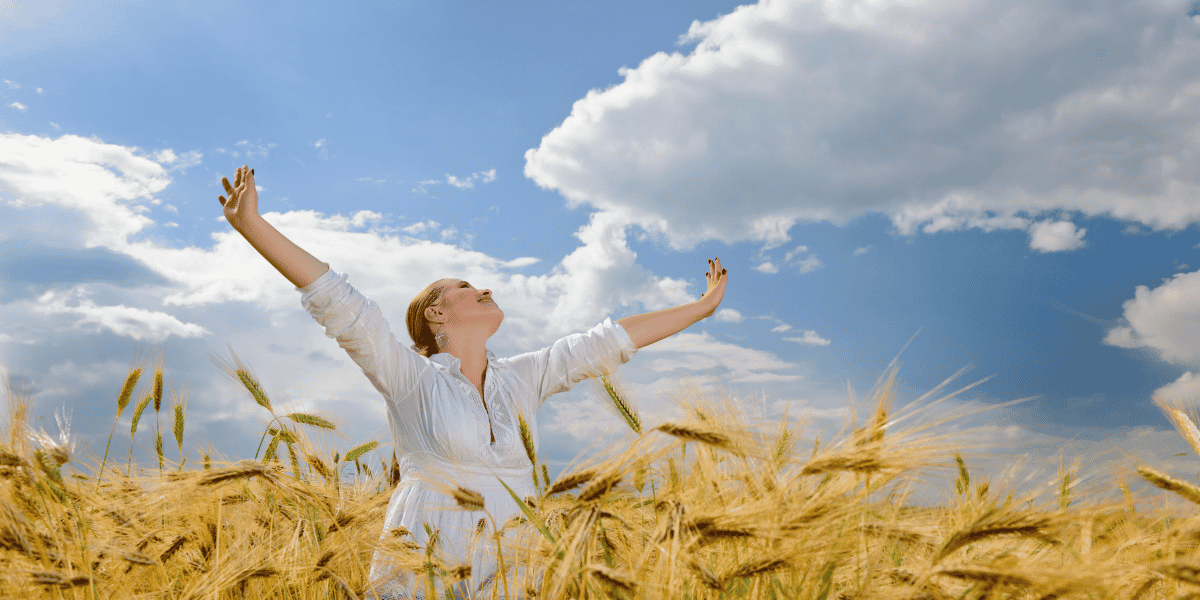 Eine Frau in einem weißen Kleid steht in einem goldenen Weizenfeld, breitet die Arme weit aus und blickt lächelnd in den Himmel. Über ihr ziehen große, helle Wolken durch einen strahlend blauen Himmel, was eine friedliche und freie Stimmung vermittelt.