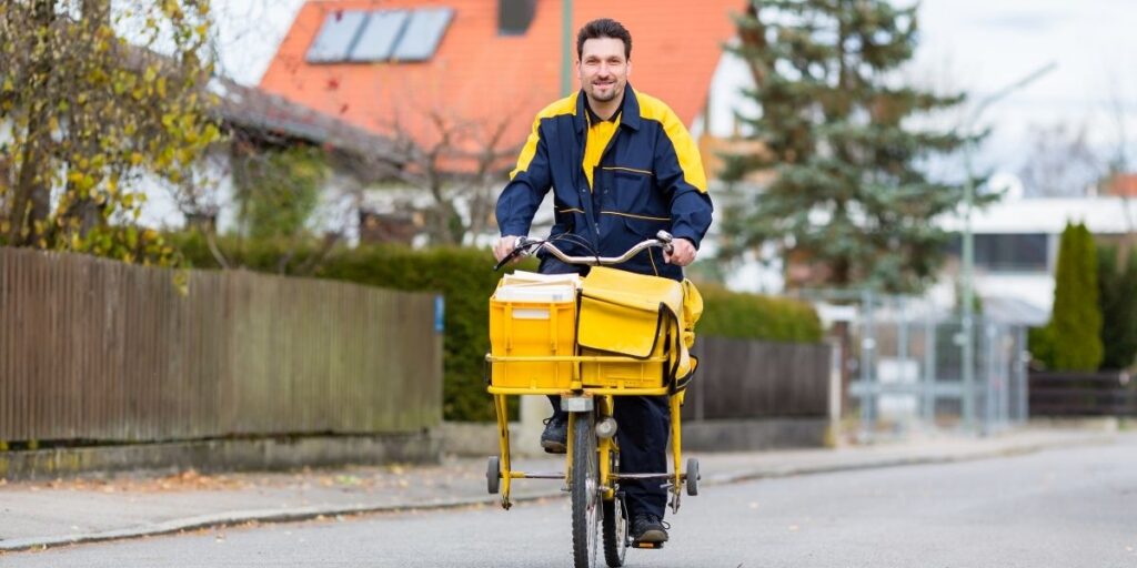 Postbote auf gelbem Fahrrad mit Transportkisten in Wohnstraße.