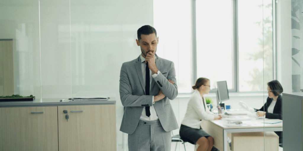 Mann im grauen Anzug steht nachdenklich in modernem Büro, zwei Personen arbeiten im Hintergrund am Schreibtisch.