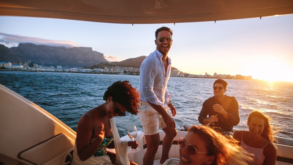 Gruppe von Menschen feiert auf einem Boot bei Sonnenuntergang vor einer Küste