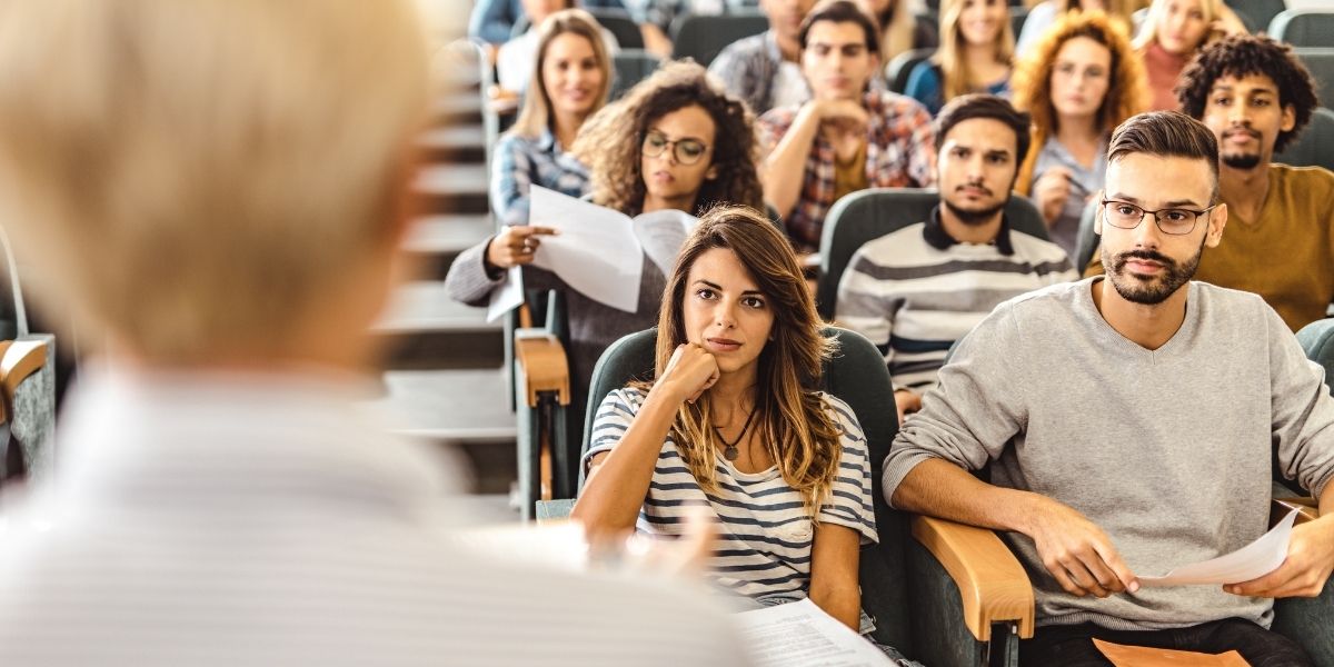 Vorlesung mit Studierenden in einem Hörsaal, Fokus auf Zuhörende und Dozent im Vordergrund.