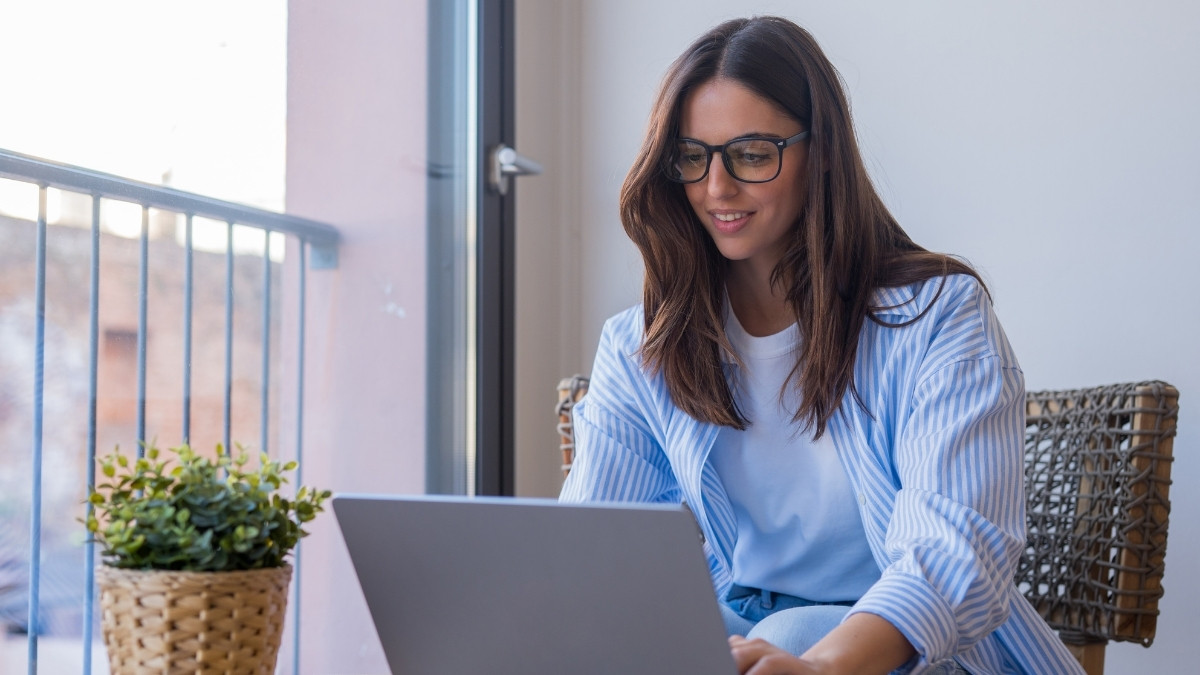 Frau mit Brille arbeitet lächelnd am Laptop neben einem Fenster