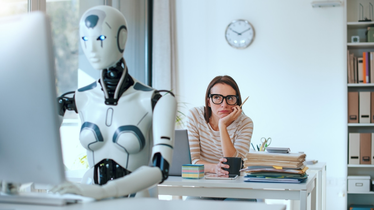 Frau sitzt nachdenklich am Schreibtisch, während ein Roboter am Computer arbeitet
